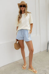 A woman stands outdoors against a white wall wearing a cream short-sleeve t-shirt, light blue drawstring shorts with white side stripes, a straw hat, sunglasses, straw sandals, and holding a woven straw bag.