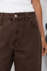 Close-up of a person wearing dark brown washed denim jeans with a white ribbed top, showing the waistband, button, belt loops, front pocket, and hand.
