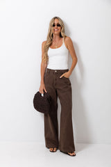 A woman modeling a pair of dark brown wide-leg jeans with a white ribbed tank top, holding a dark brown woven handbag against a plain white studio background.