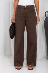 A person wearing dark brown wide-leg jeans, styled with a white top, brown sandals, and holding a dark brown woven handbag against a white draped studio backdrop.
