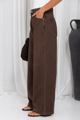 A person wearing dark brown wide-leg denim jeans, styled with a white top, black sandals, and a black crocheted bag, standing against a white curtain backdrop.