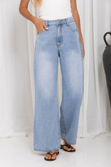 A person wearing light-wash wide-leg denim jeans with a white sleeveless top and black sandals, standing against a white draped backdrop.