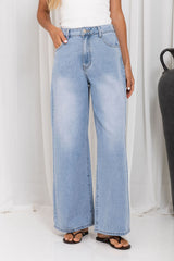 A person wearing light-wash wide-leg denim jeans with a white top and black sandals, standing against a white curtain backdrop.
