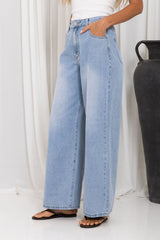 A person wearing light-wash wide-leg denim jeans with a white top and black flip-flops, standing indoors against a white curtain backdrop next to a dark vase.