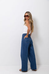 A woman modeling a pair of wide-leg blue denim jeans, paired with a white tank top and sunglasses, standing against a plain white studio background.