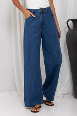 A person wearing high-waisted, wide-leg blue denim jeans with a white top and black sandals, standing against a white draped backdrop.