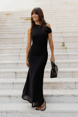 A woman wearing a black off-the-shoulder maxi dress with a sheer hem, standing on white stone steps and holding a black handbag.