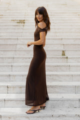 A woman wearing a dark brown off-the-shoulder maxi dress and black heeled sandals, standing on light-colored stone stairs.