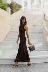 A woman wearing a sleeveless, floor-length dark brown dress walks down a grand outdoor staircase, holding a small brown patterned handbag.