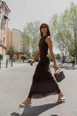 A woman walking on a city street wearing a sleeveless, dark brown, bodycon maxi dress and carrying a Louis Vuitton monogram handbag.