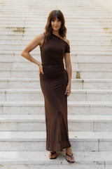 A woman wearing a dark brown one-shoulder maxi dress and brown strappy sandals, posing on a set of wide, light-colored stone stairs.