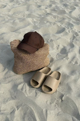 A pair of beige slide sandals and a brown baseball cap resting on a woven straw bag, all placed on a sandy beach.