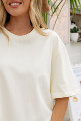 A close-up of a woman wearing a plain off-white short-sleeve crewneck t-shirt, cropped from the mouth down, with a softly blurred indoor background.
