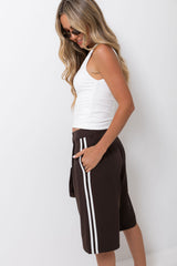 A woman wearing dark brown knee-length shorts with white side stripes, styled with a white ribbed tank top and sunglasses, standing against a plain white wall.