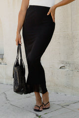 A woman wearing a black midi skirt with a sheer tulle hem, paired with a white top, black sandals, and holding a black woven handbag against a light stone wall.