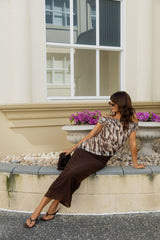 A woman wearing a brown and beige patterned top and a dark brown midi skirt sits on a stone planter ledge outdoors in front of a cream-colored building.