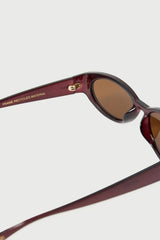 Close-up of a pair of sunglasses with a translucent burgundy frame and brown tinted lenses, showing the temple arm with 'FRAME RECYCLED MATERIAL' text and a small gold logo.