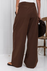 A person shown from the back wearing wide-leg brown pants, standing in a studio with a white curtain backdrop and a wooden chair partially visible on the right.