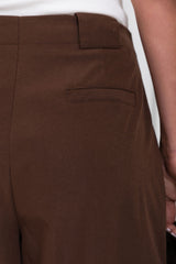 Close-up of a person wearing brown tailored trousers, showing the waistband, belt loop, front seam, and a side welt pocket. A white top is partially visible at the top edge.