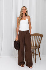 A woman stands in a studio wearing a white ribbed tank top and high-waisted, wide-leg brown trousers, holding a dark woven handbag. A wooden spindle-back chair is positioned to her right against a backdrop of white draped curtains.