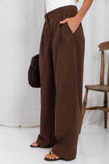 A person wearing high-waisted, wide-leg brown trousers with side pockets, styled with a white top, brown sandals, and a dark brown woven clutch bag, standing in a bright studio setting with a wooden chair in the background.