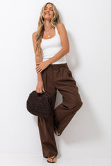 A woman posing in a white ribbed tank top and brown wide-leg trousers, holding a dark brown woven handbag against a plain white studio background.