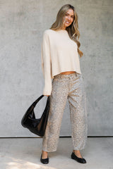 A woman stands against a textured gray wall wearing a cream knit long-sleeve crop top, beige and brown leopard-print wide-leg pants, black flats, and holding a black slouchy handbag.