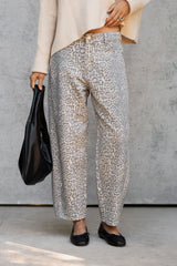 A person wearing loose, wide-leg pants with a beige and gray animal print pattern, paired with a cream sweater and black flats, holding a black handbag against a gray concrete wall.