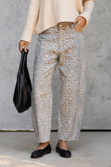 A person wearing loose, wide-leg trousers with a beige and gray animal print pattern, paired with a cream sweater and black flats, holding a black handbag against a gray concrete wall.