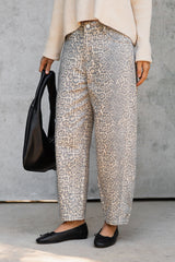 A person wearing loose, ankle-length patterned pants in beige, gray, and brown tones, styled with a cream sweater, black flats, and holding a black shoulder bag against a concrete wall.