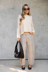 A woman stands against a textured gray wall wearing a cream knit long-sleeve top and beige-and-black leopard-print wide-leg pants, holding a black handbag.