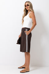 A woman modeling a pair of dark brown knee-length shorts with white side stripes, paired with a white tank top and black flip-flops, standing against a plain white studio background.