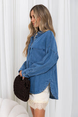 A woman wearing an oversized blue denim shirt and cream lace-trim shorts, holding a dark brown woven clutch bag, standing in front of white curtains.
