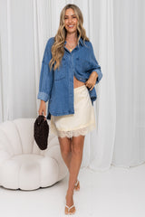 A woman models a blue denim button-up shirt and a cream lace-trimmed mini skirt, holding a dark brown textured clutch bag against a white curtain backdrop.