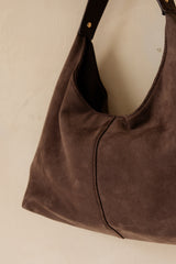 A close-up of a dark brown suede hobo shoulder bag with a black leather strap and gold-tone rivets, shown against a neutral cream-colored background.