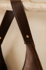 Close-up of a brown leather shoulder strap with gold-tone rivets and a suede-like brown body, hanging against a warm beige background.