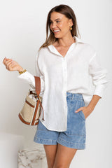 Woman wearing a white shirt and denim shorts, holding a straw bag against a plain background