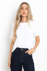 A woman with long blonde hair wearing a white short-sleeve crewneck t-shirt and dark blue jeans, standing against a plain white background.