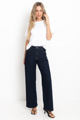 A woman modeling dark wash wide-leg jeans with a white t-shirt and black strappy heels, standing against a plain white studio background.