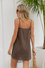 A woman wearing a brown and beige plaid mini dress with thin chain straps, shown from the back in a bright indoor setting with a potted plant.