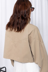 Woman wearing a stone funnel neck bomber jacket over white pants, styled with a brown suede bag, on a white background.