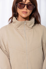 Woman wearing a stone funnel neck bomber jacket over white pants, styled with a brown suede bag, on a white background.