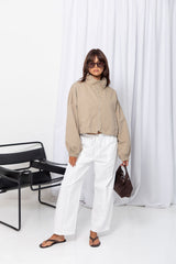 Woman wearing a stone funnel neck bomber jacket over white pants, styled with a brown suede bag, on a white background.