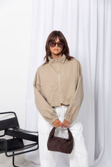 Woman wearing a stone funnel neck bomber jacket over white pants, styled with a brown suede bag, on a white background.
