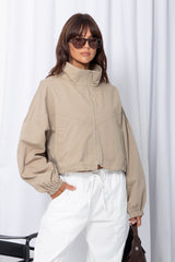Woman wearing a stone funnel neck bomber jacket over white pants, styled with a brown suede bag, on a white background.
