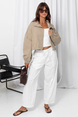 Woman wearing a stone funnel neck bomber jacket over white pants, styled with a brown suede bag, on a white background.