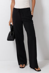A model wearing black wide-leg drawstring pants, styled with a white top, black sandals, and holding a black handbag against a plain white studio background.
