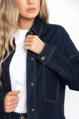 A close-up of a woman wearing a dark indigo denim button-up shirt jacket over a white t-shirt, with her hand adjusting the collar.