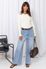 Woman wearing a white long-sleeve top with light-wash wide-leg jeans and a brown belt, carrying a brown handbag, on a white background.