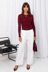 Woman wearing a burgundy long-sleeve top with white drawstring pants, carrying a burgundy bag, on a white background.